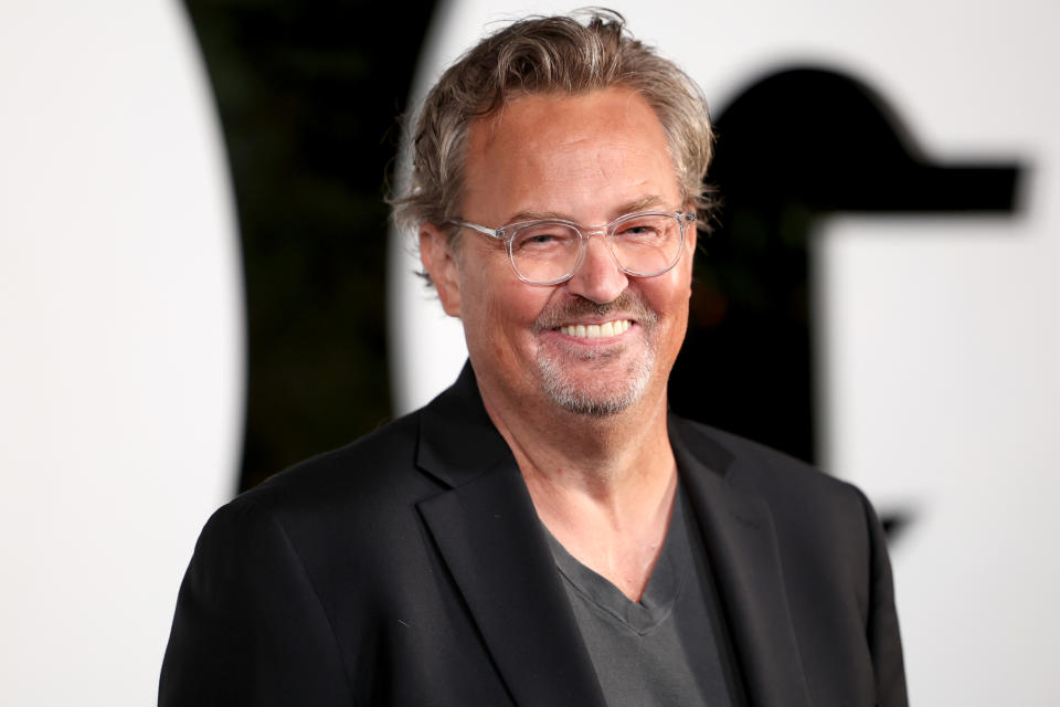 Matthew Perry smiles while wearing glasses, a black blazer, and a dark V-neck shirt