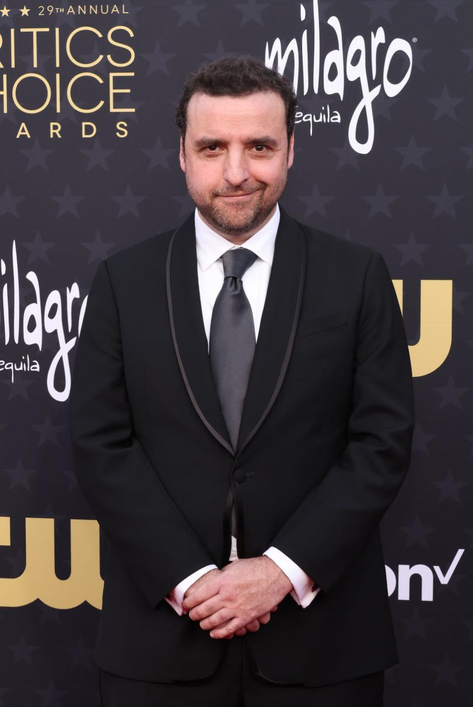 david krumholtz at the 29th annual critics choice awards 2024 he is wearing a black suit and grey tie, and standing with his hands crossed as he smiles at the camera