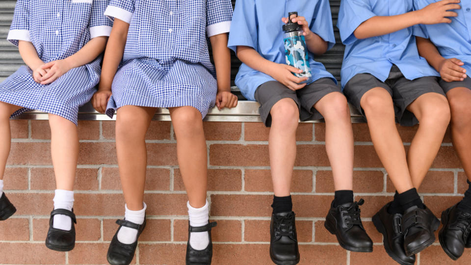 Low section view of five school children sitting on brick wall wearing school uniform. Children's legs and feet in black shoes hanging down