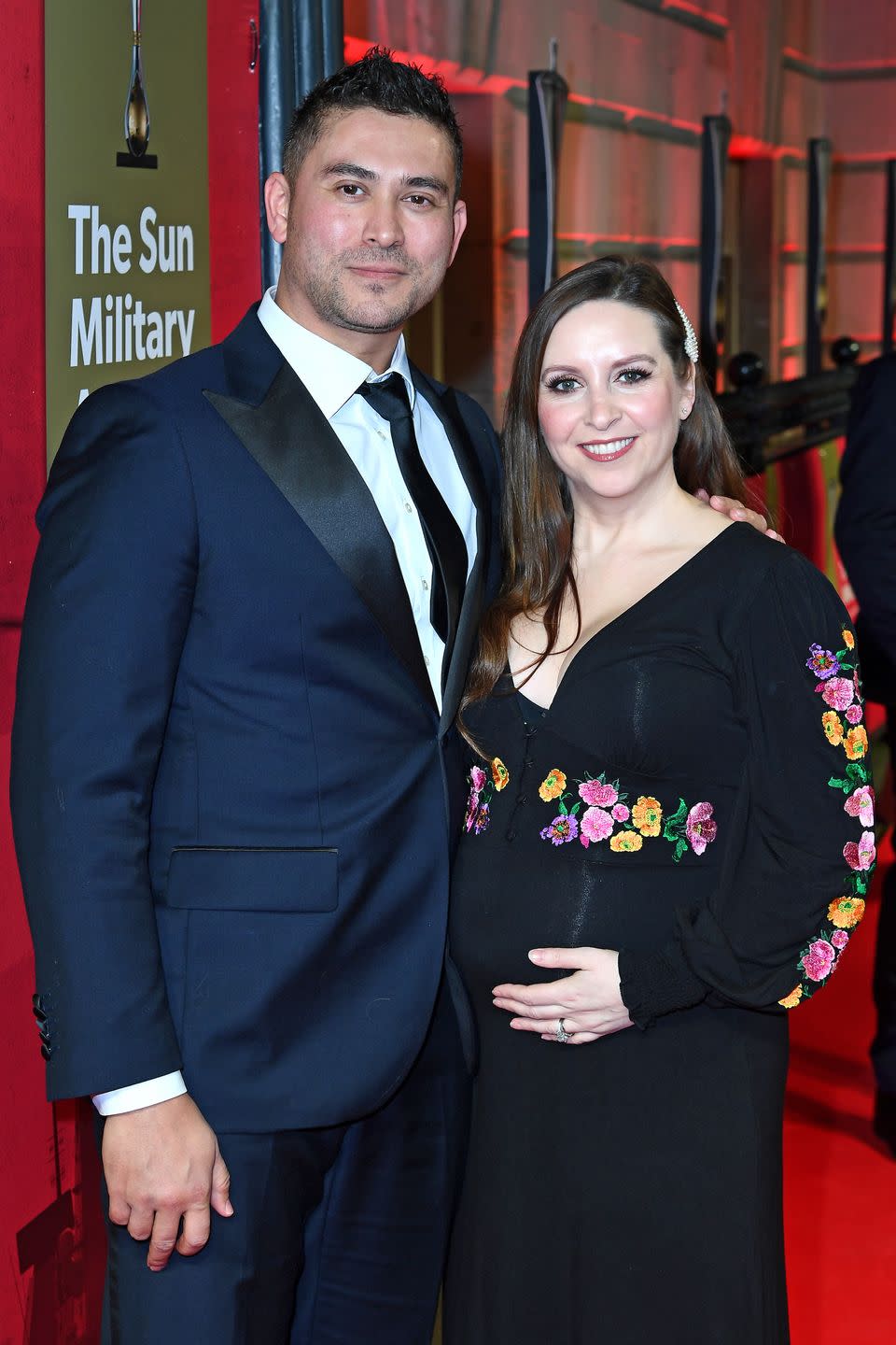 rav wilding and jill morgan, a man and woman stand next to each other smiling at the camera, he wears a navy suit and she wears a black dress as she cradles a baby bump