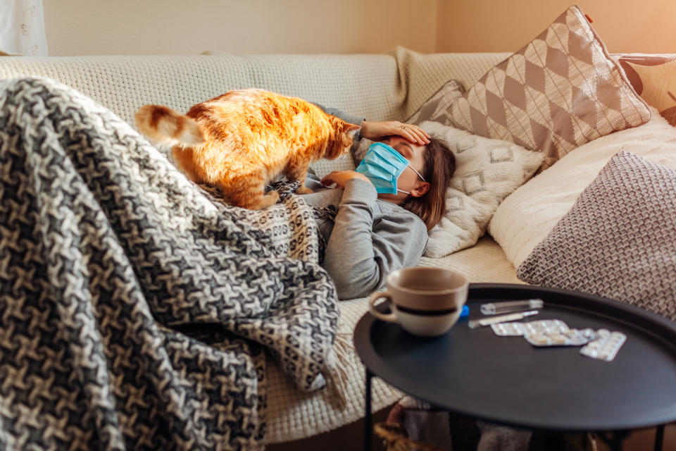 Sick woman having flu or cold. Girl lying in bed with cat having headache wearing protective mask by pills and water on table.