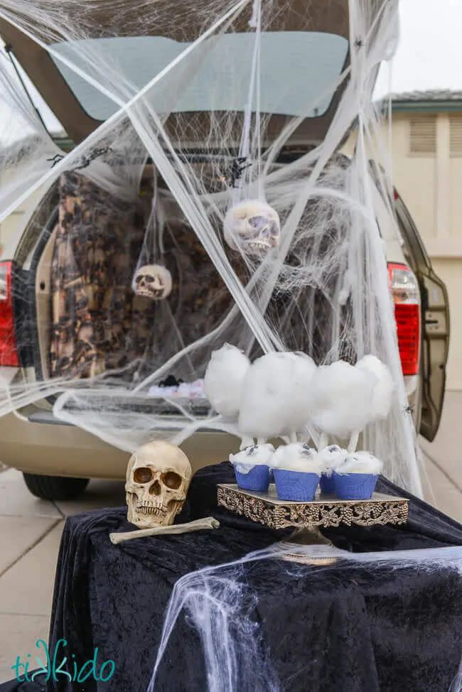 spider den trunk or treat with open tailgate of vehicle filled with faux spider webs, spiders and twh sitting in front of it with a skull, more webs and a tray of spider web cotton candy
