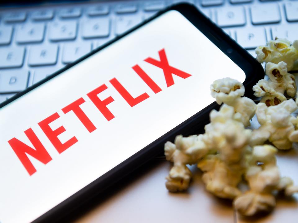 The Netflix logo is displayed on a smartphone with popcorn and laptop keyboard in the background.