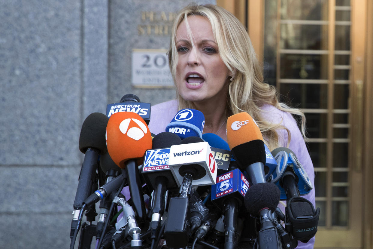 Stormy Daniels speaks in front of a bank of microphones, with a glass-partitioned door behind her.
