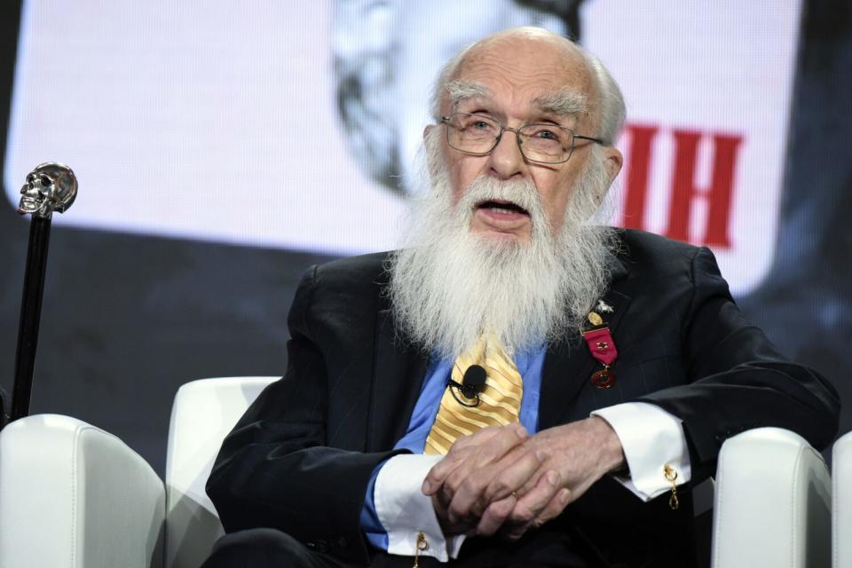 A man with a long white beard and glasses sits in a chair onstage.