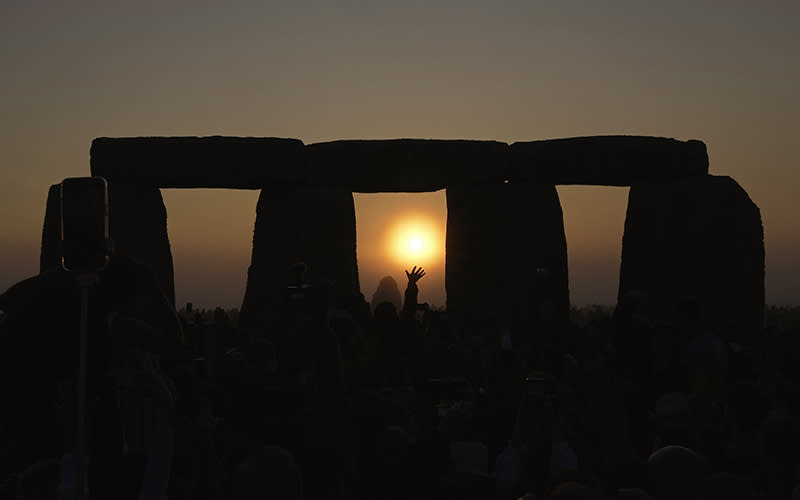 The sun sets at Stonehenge during the summer solstice. The sky is dimming and the sun is framed in one of the openings in Stonehenge, while the silhouette of a reveler's hand is seen reaching up toward the sun.