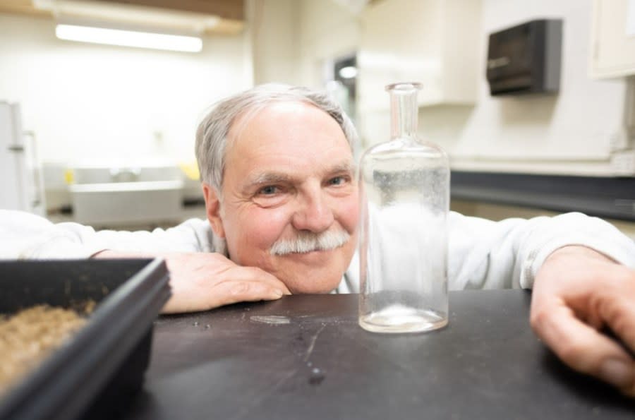 Frank Telewski, with short gray hair and a mustache, bends down to eye level with a small glass bottle.