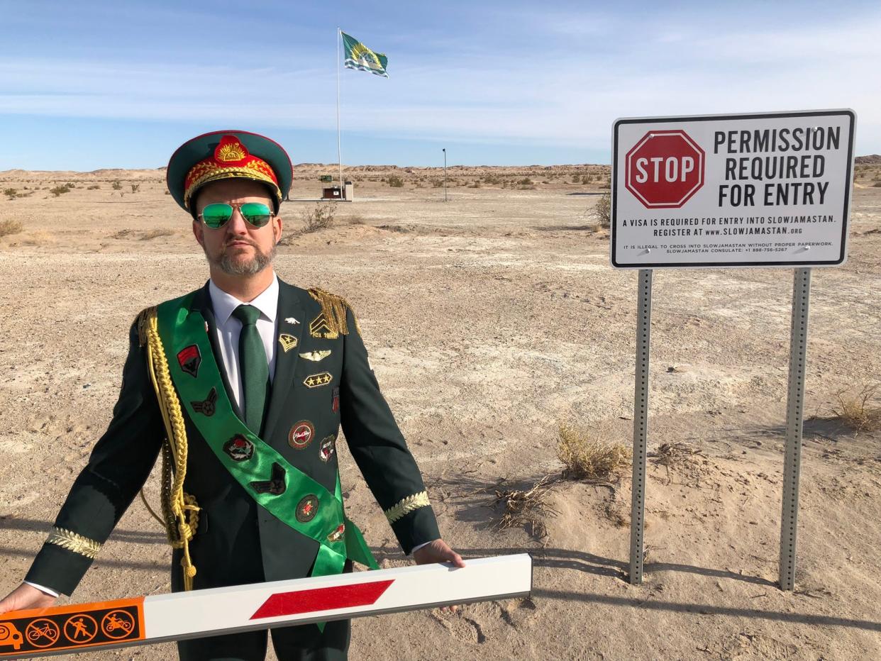 Randy Williams standing behind a barrier leading to the micronation of Slowjamastan while wearing an official green uniform.