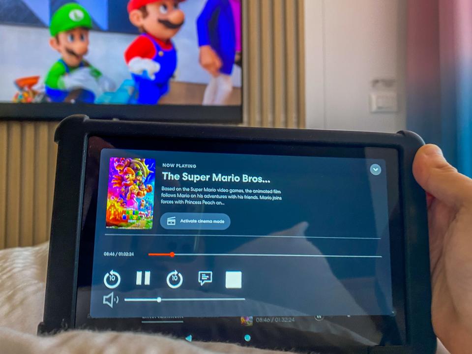 A hand holds a black tablet with Mario and Luigi on the TV behind it.