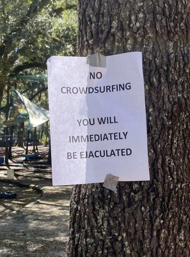 Sign on tree reads: "NO CROWDSURFING YOU WILL IMMEDIATELY BE EJACULATED" with a playground in the background