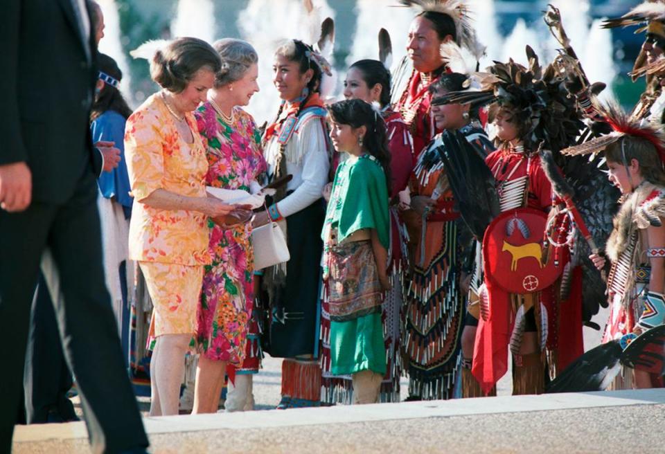 Queen Elizabeth smiles at a group of people dressed in traditional Native American attire, representing the Texas American Heritage Society, after accepting a gift from the group outside of the Hall of State in downtown Dallas.