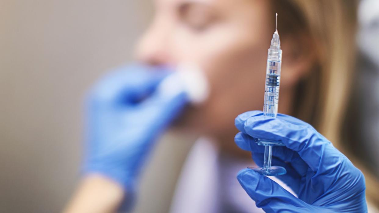  Close up on a syringe in a gloved hand. A woman sitting in a chair can be seen blurred in the background, with the provider's other hand holding a cotton swab to her face. 