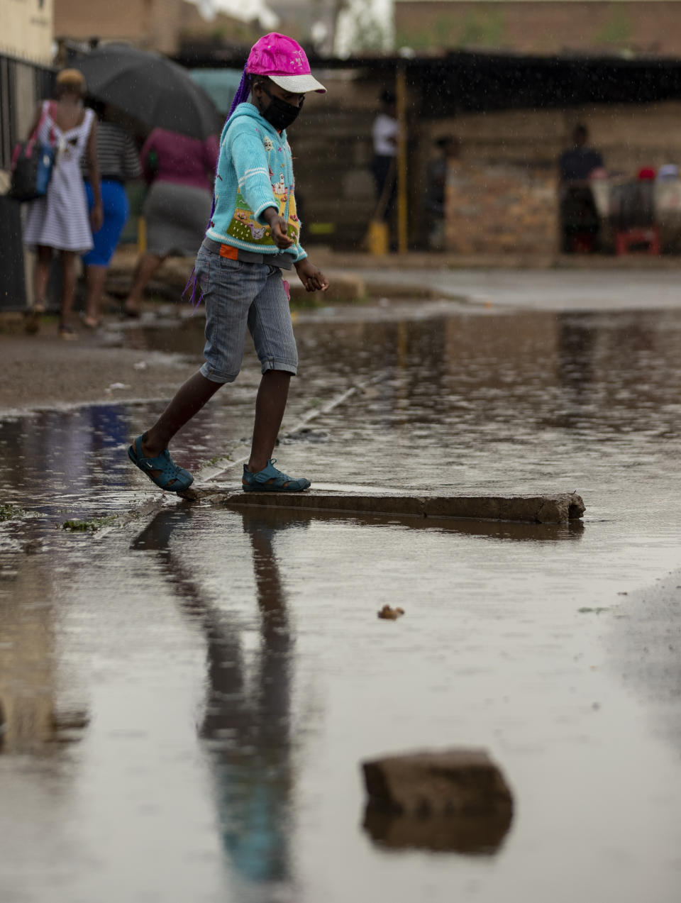 A young girl wearing a mask to help protect themself from the coronavirus crosses a waterlogged street after rainfall in Thokoza, east of Johannesburg, South Africa, Thursday, Jan. 14, 2021. South Africa is struggling to cope with a spike in COVID-19 cases that has already overwhelmed some hospitals, as people returning from widespread holiday travel speed the country's more infectious coronavirus variant. (AP Photo/Themba Hadebe)