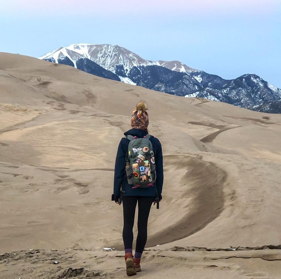 Emily, wearing a black hoodie and leggings, boots, a backpack covered in patches, and a winter hat, faces sand dunes and mountains.