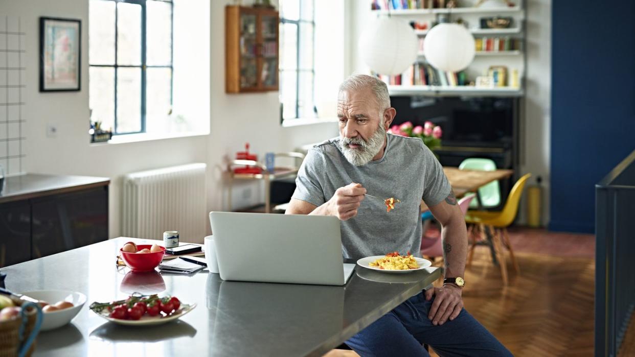 senior man sitting in kitchen eating breakfast and looking at laptop