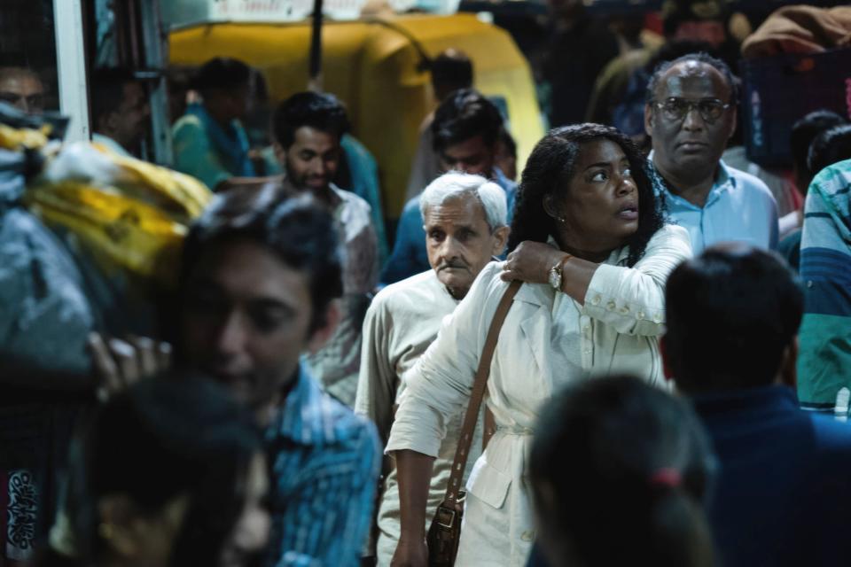 Aunjanue Ellis-Taylor in the foreground looking back in a crowded street scene, with onlookers and a tuk-tuk in the background in a scene from "Origin"