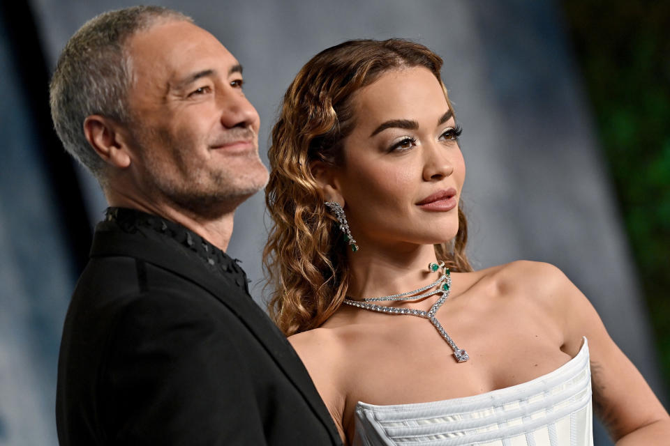 A close-up of Taika Waititi and Rita Ora posing together on the red carpet, he's in a black suit and she's in a white strapless gown