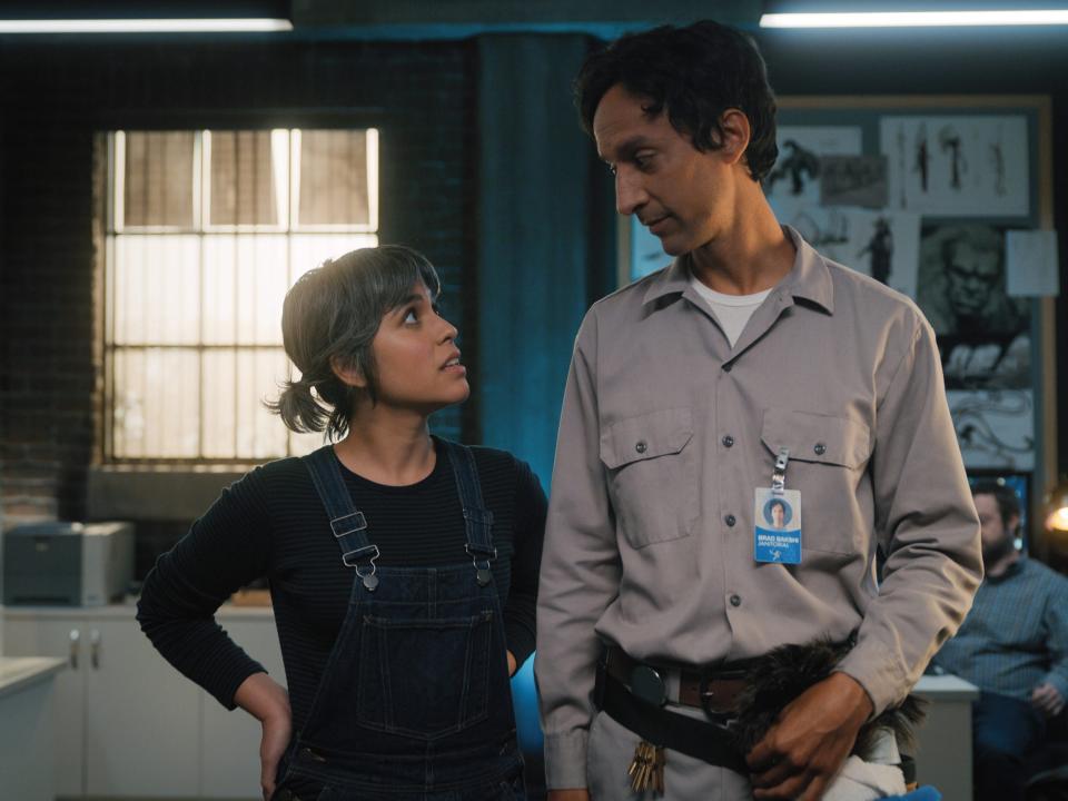 ashly burch and danny pudi as rachel and brad in mythic quest. they're standing in a dimly lit room with some computers and desks in it, looking at each other intently. ashly is wearing a black shirt and overalls, her hair in pigtails. danny is in a beige janitorial jumpsuit