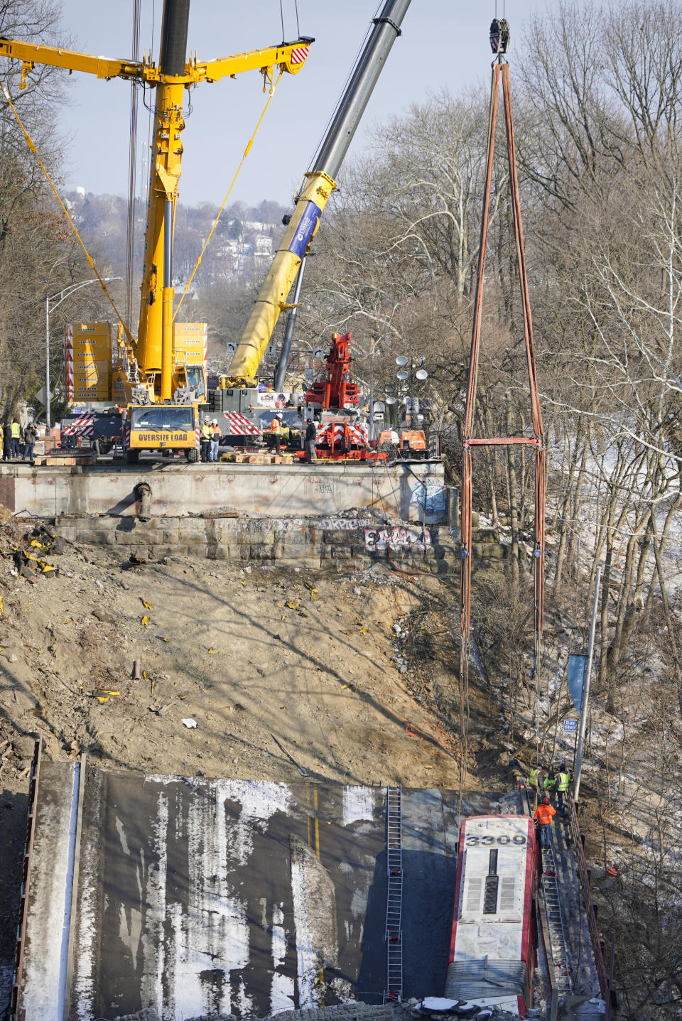 Workers prepare the bus, lower right, that was on a bridge when it collapsed Friday as they work to remove it with a crane during the recovery process on Monday Jan. 31, 2022 in Pittsburgh's East End. (AP Photo/Gene J. Puskar)