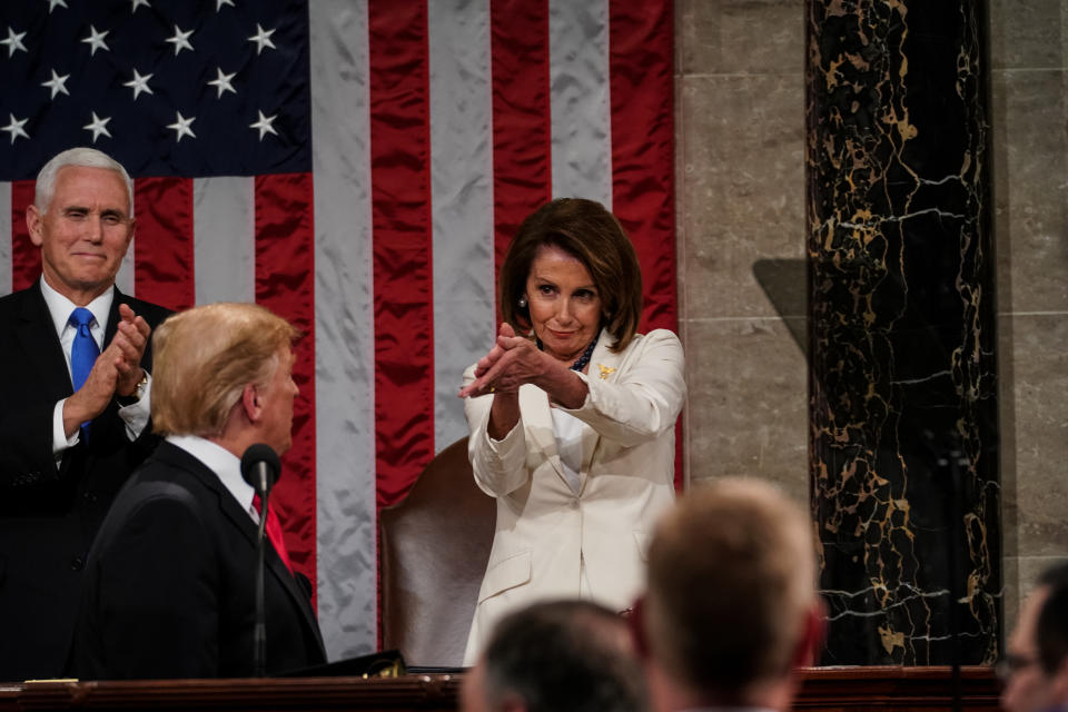 Mike Pence, Donald Trump, and Nancy Pelosi at a public event, with Pelosi applauding while Trump stands and Pence claps in the background near an American flag