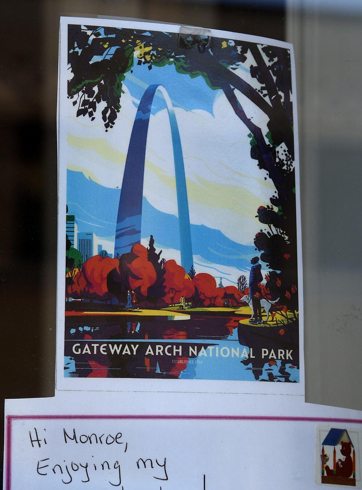 Little Brown Bear Adventures postcard on a downtown Monroe store front, above Gateway Arch National Park.