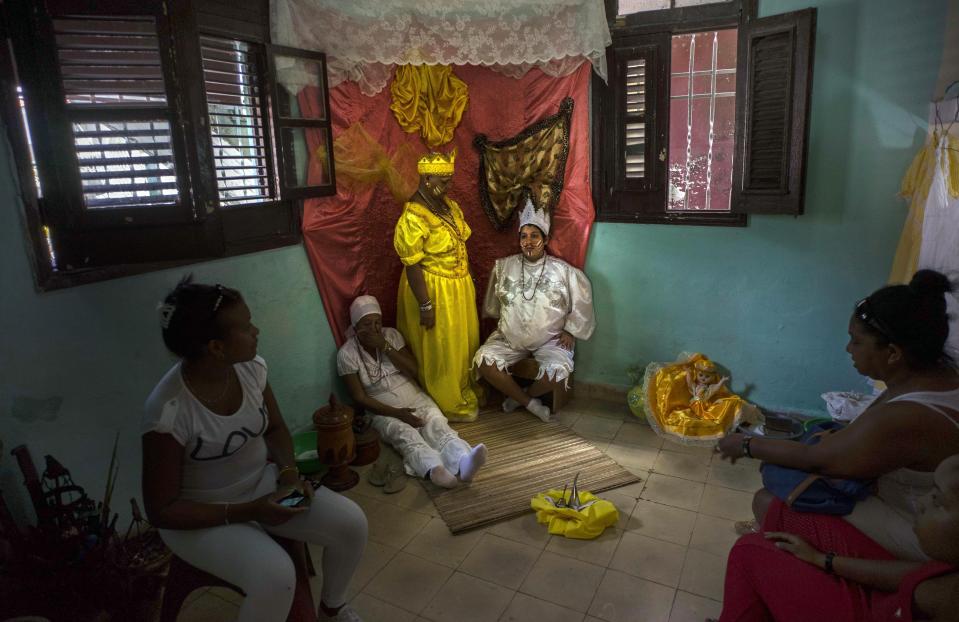 FILE - Marisa Ramirez Gutierrez, standing center, holds still on her symbolic throne dressed in the robe of the the "Iyawo," or bride, alongside a pregnant woman who represents birth, and her mother, who represents a woman who has already given birth, inside her home as part of many initiation ceremonies marking the start of her one-year journey to become a Yoruba priestess in Havana, Cuba, Sept. 11, 2015. Santería was born as a form of quiet resistance among Cuba’s Black communities. (AP Photo/Ramon Espinosa, File)