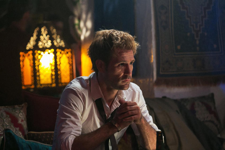 Matt Ryan as John Constantine, sits contemplatively in a dimly lit room decorated with ornate objects and fabrics