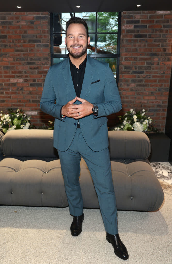 Man in tailored suit posing with hands together, smiling at a formal event