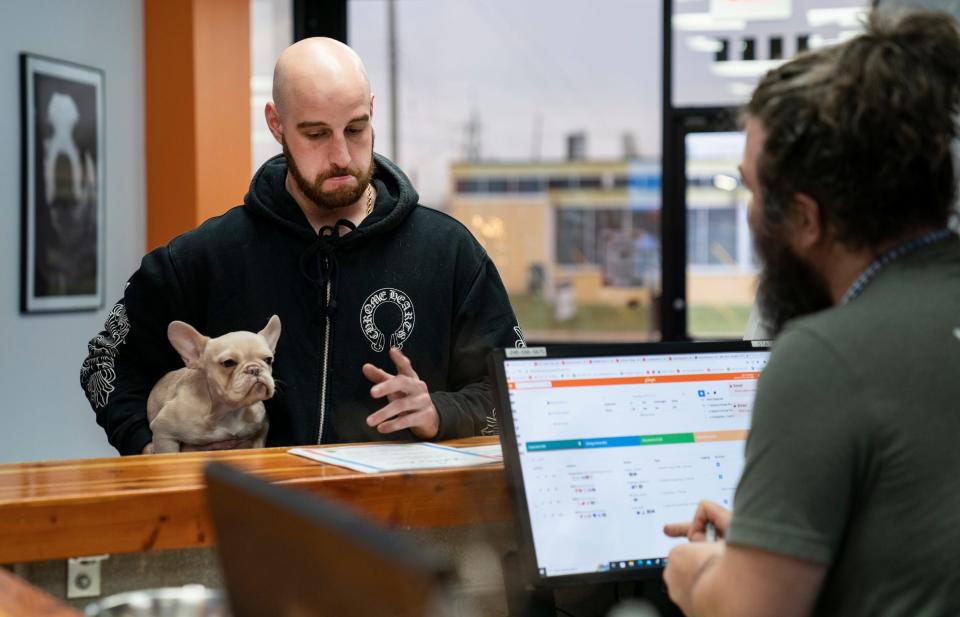 Jordon Machlis, 23, of Novi, left, holds his 7-month-old French Bulldog Bella as he checks her in with John Spreitzer, 32, the front desk greeter at Canine To Five on Dec. 22, 2022. Canine To Five, a boarding and dog daycare facility, has had to raise their prices to retain trained staffers and to cover the inflation of running the facilities and dog supplies.