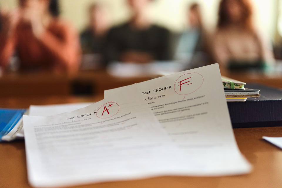 Papers on a desk with "A+" and "F" grades, students in the blurry background