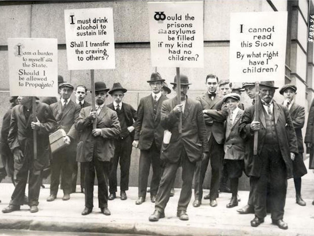 A black and white photo shows men carrying signs reading eugenic slogans like ""I am a burden to myself and the State. Should I be allowed to propagate?," "I must drink alcohol to sustain life. Shall I transfer the craving to others?," "Would prisons and asylums be filled if my kind had no children?," and "I cannot read this Sign. By what right have I children?"