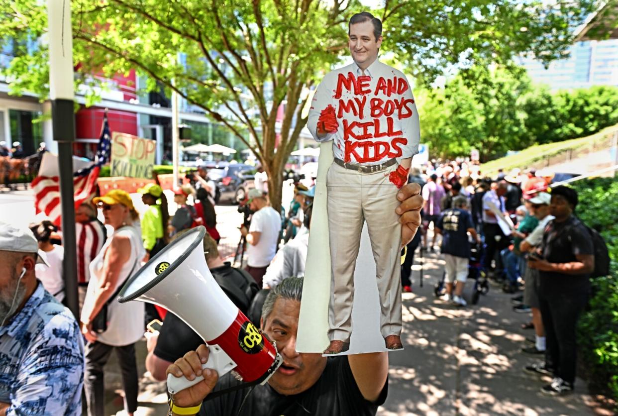 A crowd of protesters, one with a megaphone and a cutout of Ted Cruz with red hands and the words "Me and my boys kill kids."