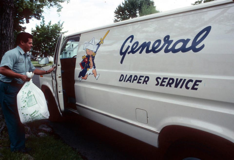 A man wearing a uniform loads a bag into a General Diaper Service van, which features a cartoon child in a sailor outfit with a diaper pin