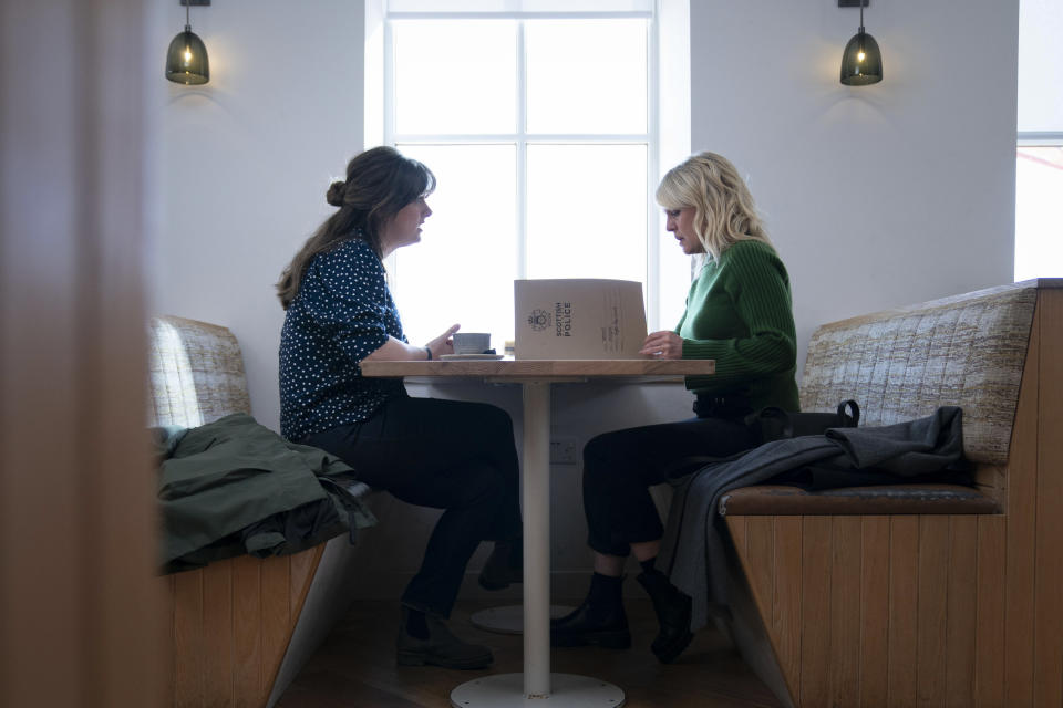 Tosh (Alison O'Donnell) and Calder (Ashley Jensen) sit at a booth in a brightly lit cafe. Tosh is talking to Calder, who has a police dossier open on the table in front of her and is reading it