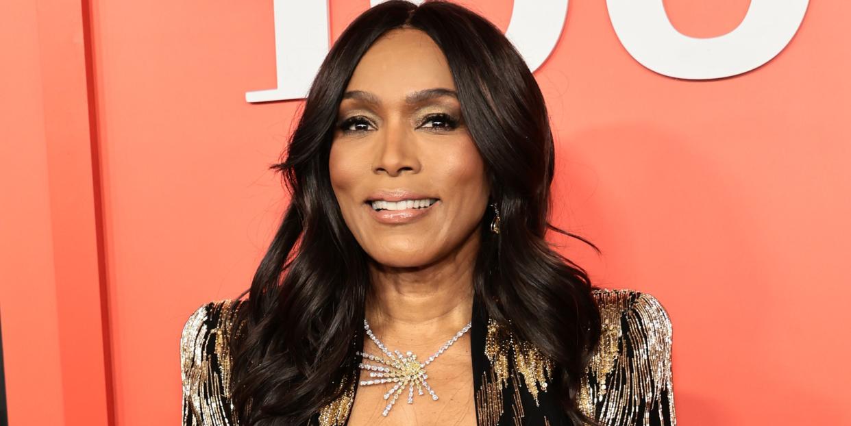 angela bassett, a woman stands looking at the camera and smiling, she has dark long hair worn down loose, wears a sparkly gold jacket and trouser suit