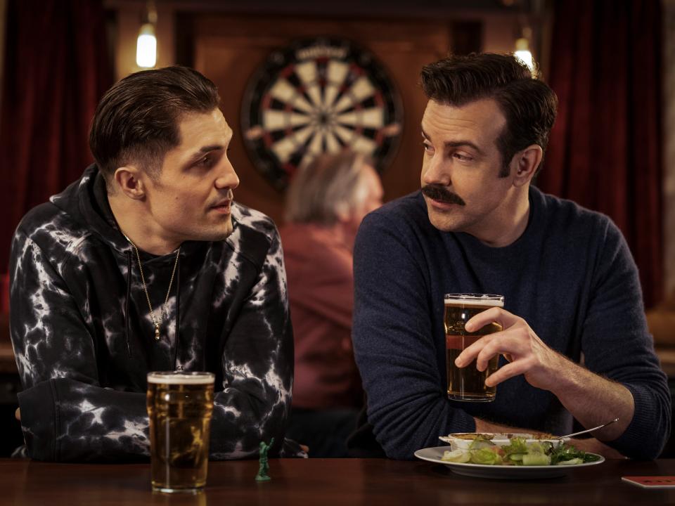 Phil Dunster as Jamie Tartt and Jason Sudeikis as Ted Lasso sit at a bar with beers on "Ted Lasso."