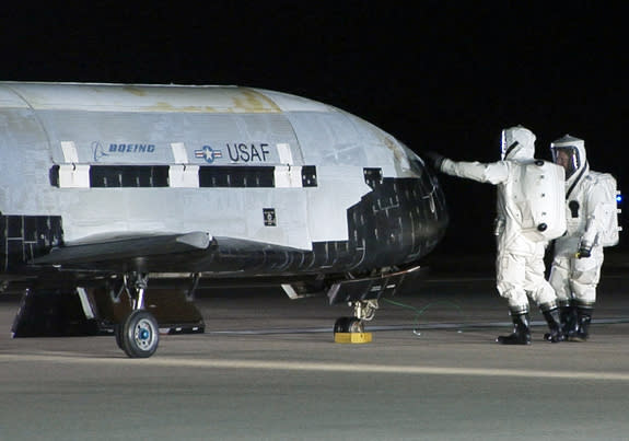 An X-37B robotic space plane sits on the Vandenberg Air Force base runway during post-landing operations on Dec. 3, 2010. Personnel in self-contained protective atmospheric suits conduct initial checks on the robot space vehicle after its landi