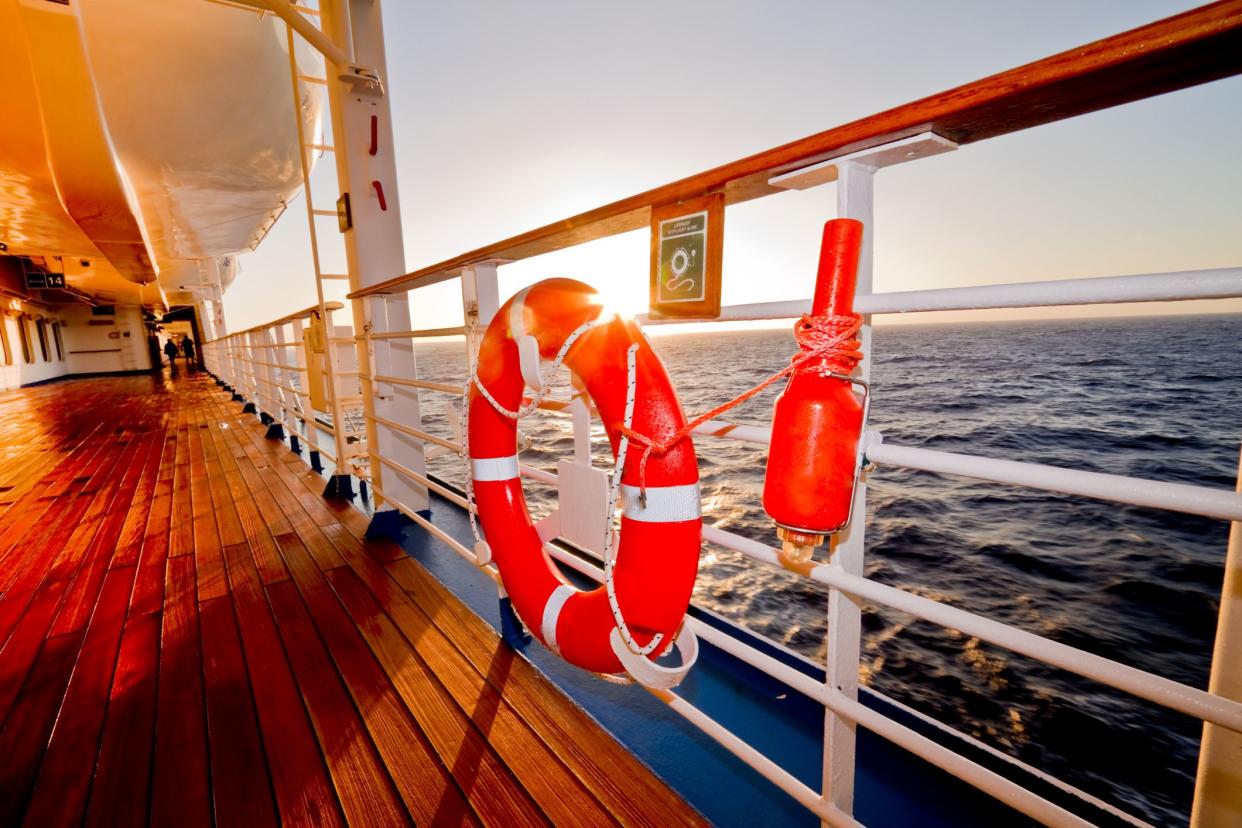 Hanging orange safety beacon and life preserver ring on the railing of a wooden promenade deck of a cruise ship during dusk, ocean in the background