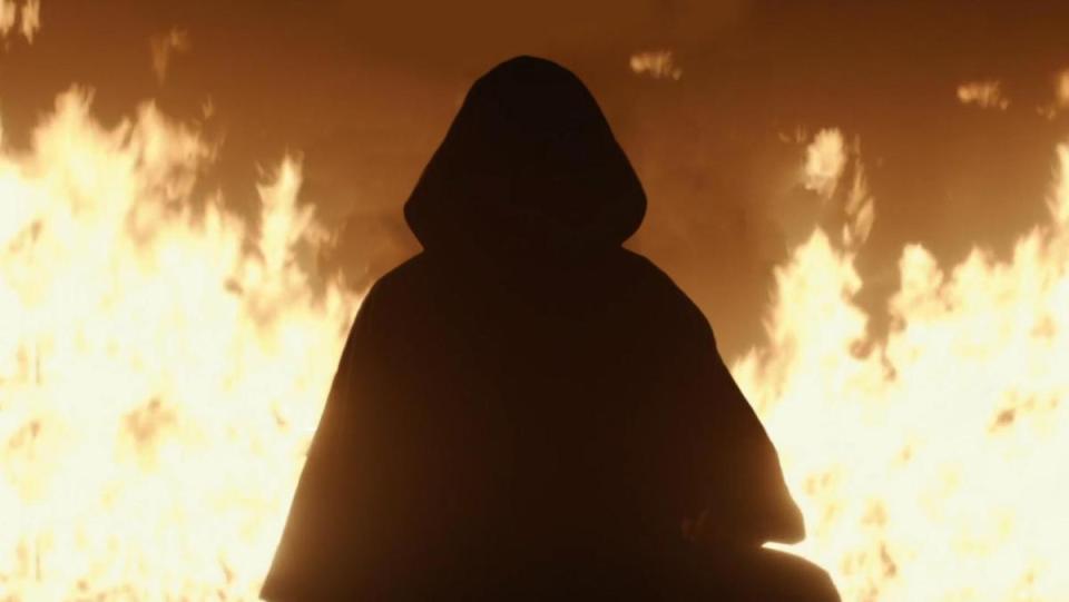 A hooded figure stands in front of flames in Loki