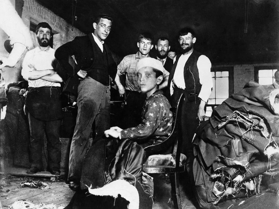 A group of men and a young boy are photographed inside a small New York City sweatshop, circa 1880s.