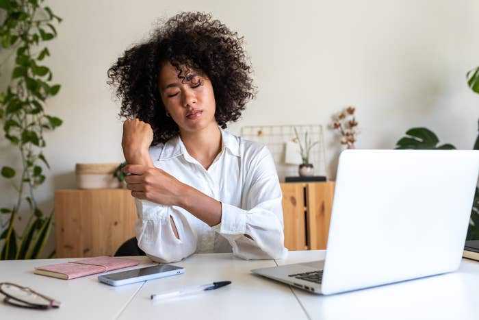 Woman at desk rubbing sore wrist, sitting by a laptop, notebook, and smartphone, with plants in the background