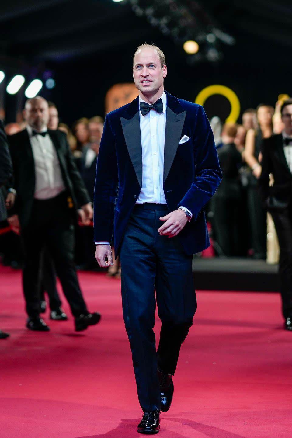 prince william walks on a red carpet while looking slightly up and smiling, he wears a blue velget suit with a bow tie and white collared shirt