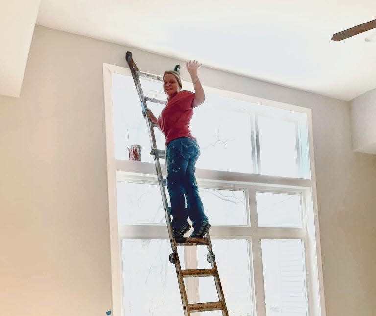 Image of Melissa Angle balancing on a ladder, waving with her left hand. She has blonde hair that's pulled back and wears a red shirt and blue denim jeans spattered with paint. Behind her is a wide paneled window with a bucket of paint resting on the upper sill, where is the sun is shining brightly. 