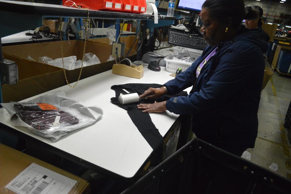 A woman in a dark hoodie lint rolls a black sweater on a brightly lit table inside a warehouse.