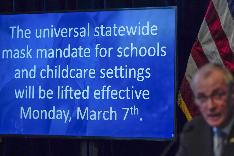 New Jersey Gov. Phil Murphy speaking in front of a large screen, on which read the announcement about lifting the mask mandate.