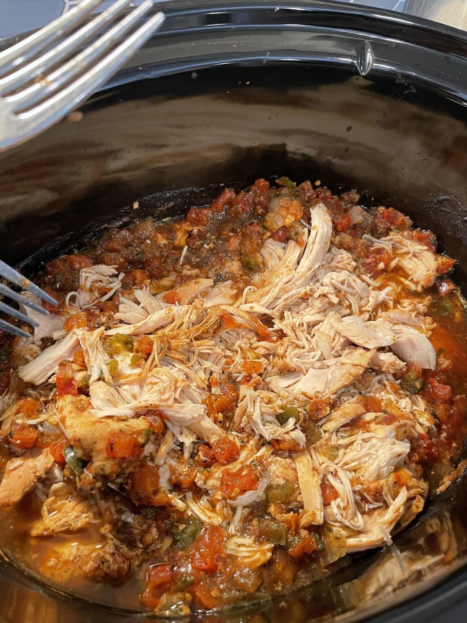 Shredded chicken cooking in a slow cooker with a tomato-based sauce and herbs. Two forks are visible in the image