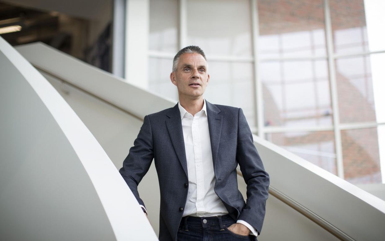 Tim Davie stands on a staircase wearing a suit and white shirt, with no tie