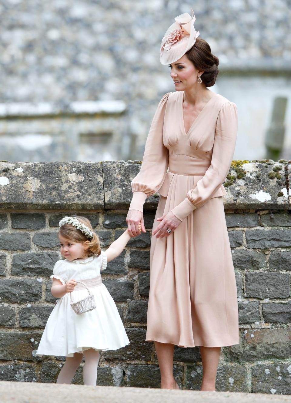 Princess Charlotte and Kate Middleton stand in front of a stone wall.