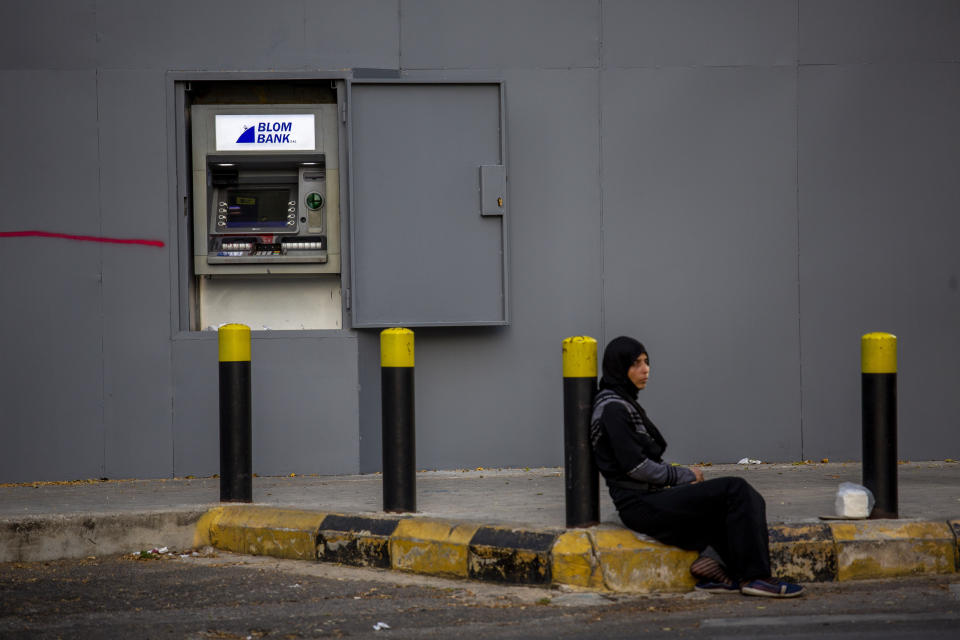 In this Thursday, May 21, 2020 photo, a woman begs on a sidewalk in front an ATM machine covered by iron shields along the facade of a bank to prevent acts of sabotage, in Beirut, Lebanon.Riad Salameh, Lebanon’s long-serving central bank governor, was touted as the guardian of Lebanon’s monetary stability as he steered the tiny country's finances through post-war recovery and various bouts of unrest for nearly three decades. Now he is being called a “thief” by some protesters, who accuse him of being part of the ruling elite whose corruption and mismanagement has driven the country to the edge of bankruptcy. (AP Photo/Hassan Ammar)