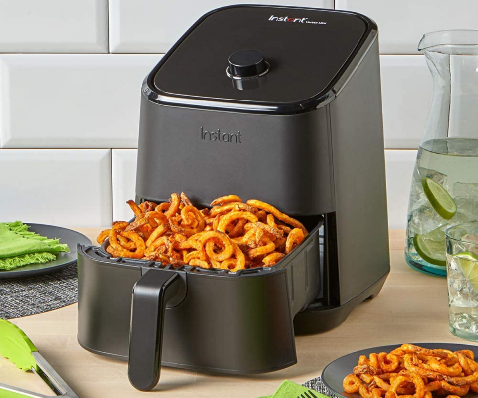 The Instant Pot Air Fryer in black sits on a wooden kitchen bench infront of white tiles with the black bottom tray open showing crispt curly sweet potato fries.